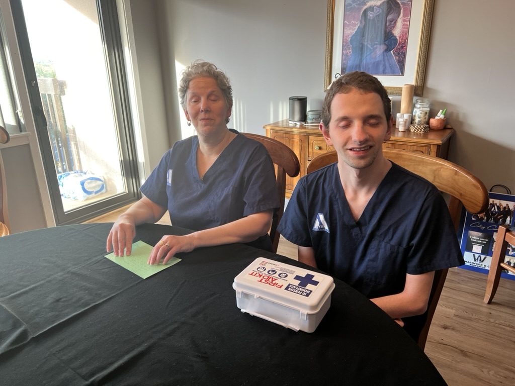 Christine, sits next to Braden at a table. On the table Christine has a notes card in braille and Braden has a first aid kit in front of him.
