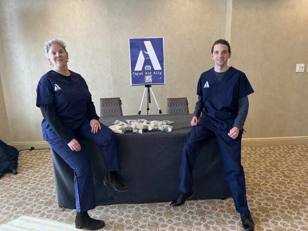 First Aid Ally's co-owners Christine and Braden, both sit at opposite ends on the edge of a 6 foot table. In the center are first aid kits. Christine and Braden are dressed in navy blue scrubs with the first aid ally logo on the shirt.