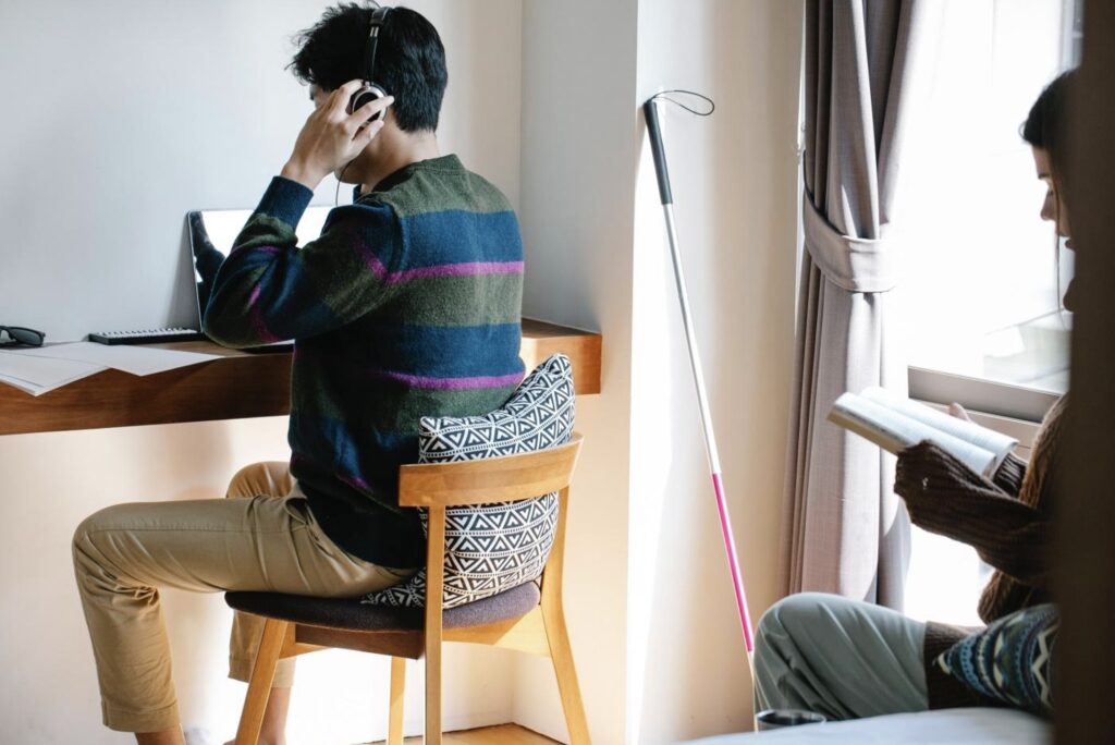 Two students study. One at a desk and one sitting on the edge of a bed. There is a white cane leaning on the wall near the student at the desk.