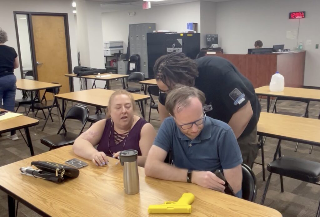 A firearm safety instructor leans in to speak to a woman with dirty blond hair in a ponytail. On the desk folded is a white cane.