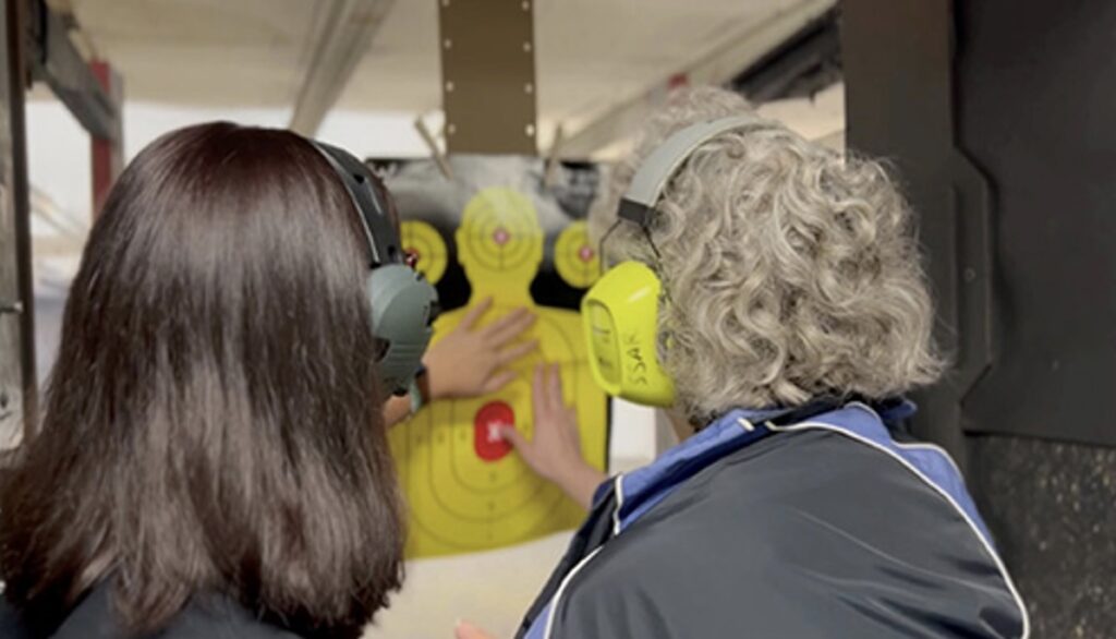 An instructor and her student, stand in a gun range booth. Getting familiar with a yellow target with a red bullseye.