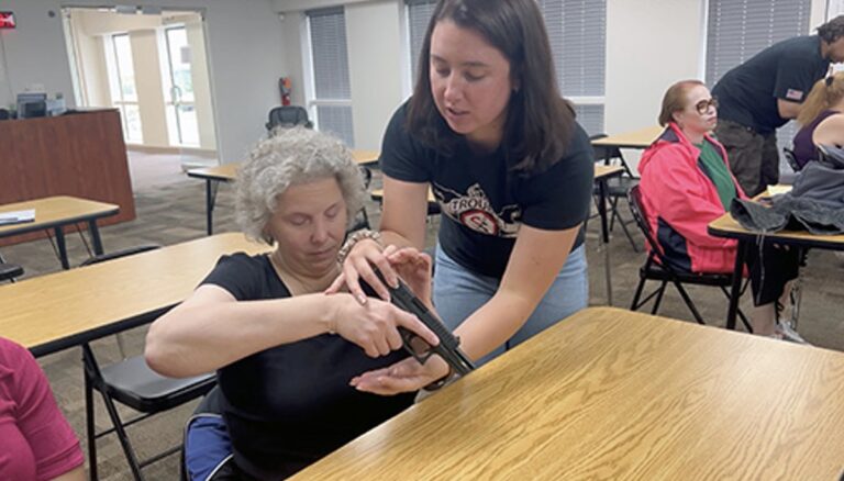 A firearms safety instructor stands over and guides a blind woman to get acquainted with a firearm.