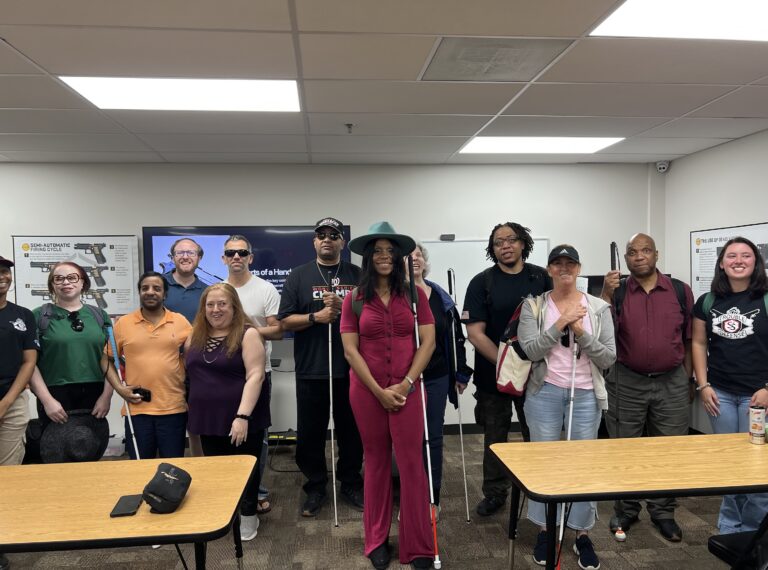A group of blind and low vision students stand in between their firearms safety instructors. Some students with white canes. All are smiling.